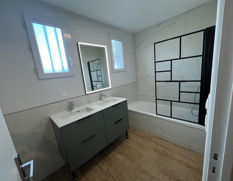 Rénovation salle de bain moderne – AC2A Aubance Salle de bain rénovée avec double vasque, miroir lumineux et pare-baignoire noir, réalisée par AC2A Aubance.